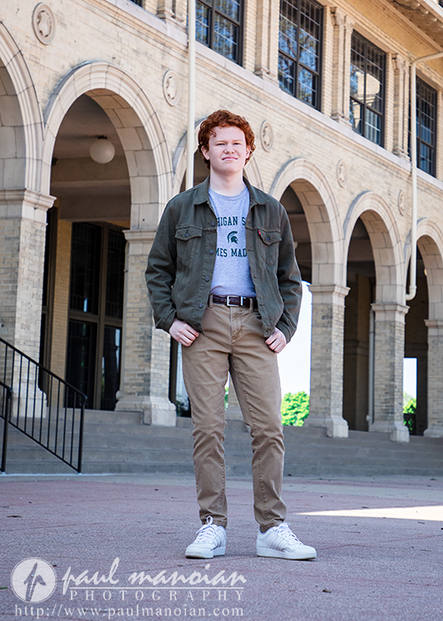 High school senior boy  with red hair stands confidently in front of a brick building with large arches and windows, capturing striking Senior Pictures for Utica Academy of International Studies in Detroit.