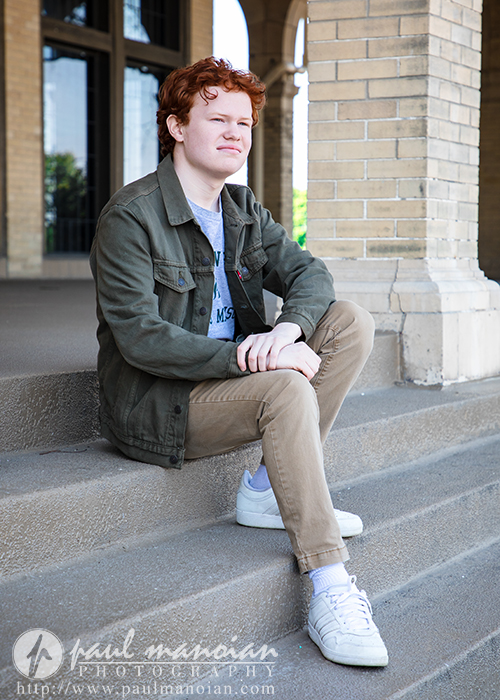 High school senior boy sits on outdoor steps in Detroit, wearing a green jacket, khaki pants, and white sneakers—perfect for unique Senior Pictures at Utica Academy of International Studies.