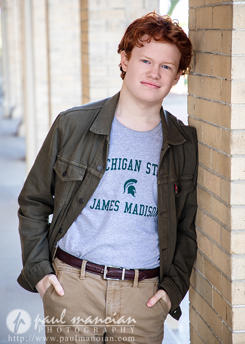 High school senior boy with red hair leans against a brick wall, wearing an olive jacket and Michigan State t-shirt, smiling during senior pictures for Utica Academy of International Studies.