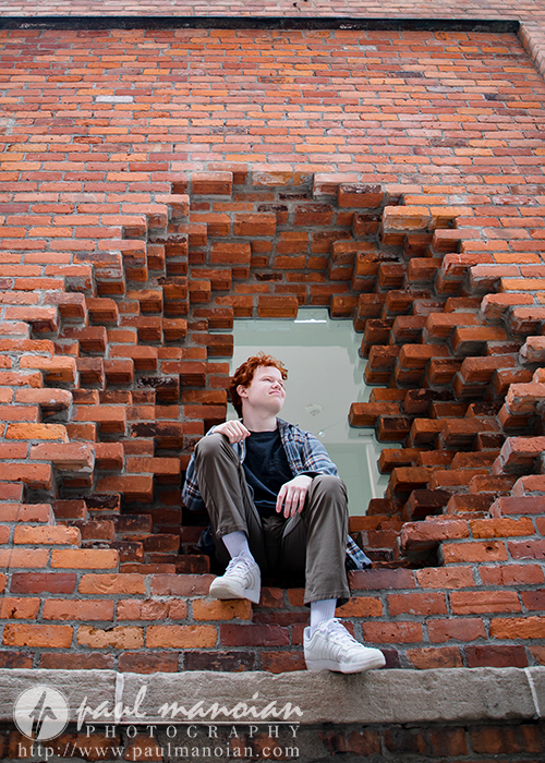 High school senior boy sits in a circular brick window, looking to the side, with a brick wall background—perfect for unique Senior Pictures at Detroit’s Utica Academy of International Studies.