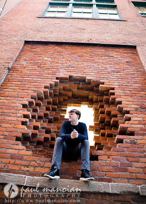 A high school senior sits in a circular hole in a brick wall, looking to the side, with a window above—perfect for unique Notre Dame Prep senior pictures or a sunrise session in Detroit.
