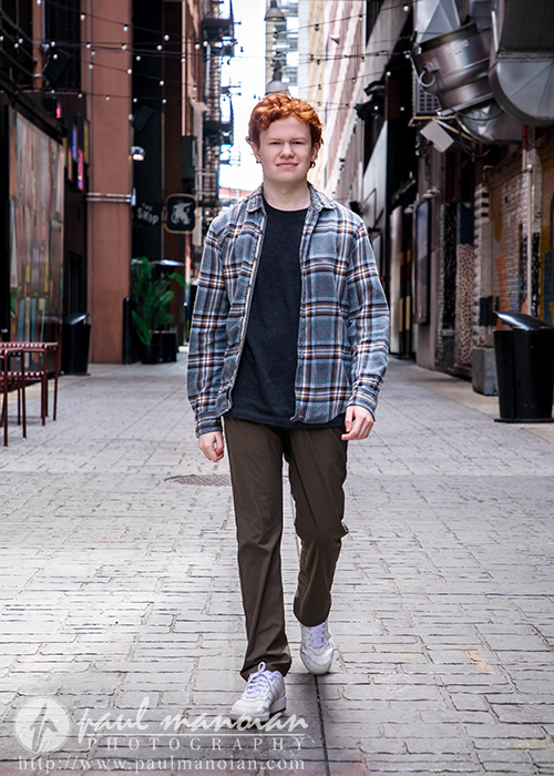 High school senior with red hair in a plaid shirt walks confidently down a brick alley lined with buildings and string lights, capturing unique senior pictures for Utica Academy of International Studies in vibrant downtown Detroit.