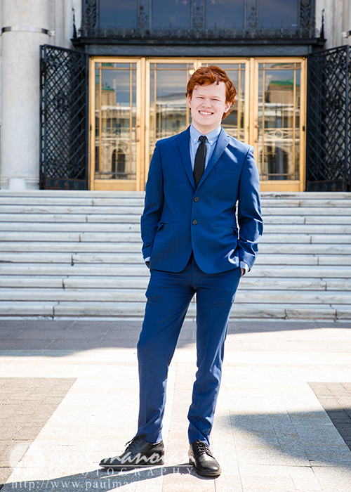 A high school senior in a blue suit stands smiling in front of gold doors and stone steps, celebrating his Senior Pictures for Utica Academy of International Studies in Detroit.