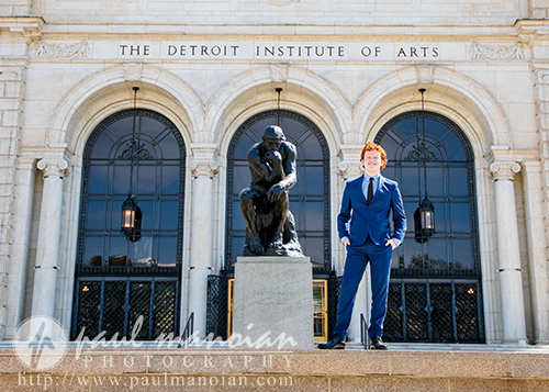 A high school senior in a blue suit stands next to The Thinker statue outside the Detroit Institute of Arts, capturing memorable Senior Pictures for Utica Academy of International Studies.