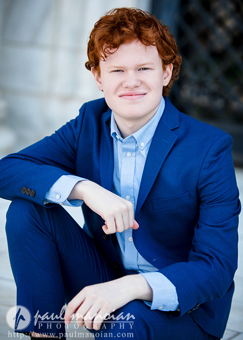 High school senior with red hair in a blue suit sits outdoors in Detroit, smiling at the camera with one hand resting on their knee—perfect for Senior Pictures at Utica Academy of International Studies.