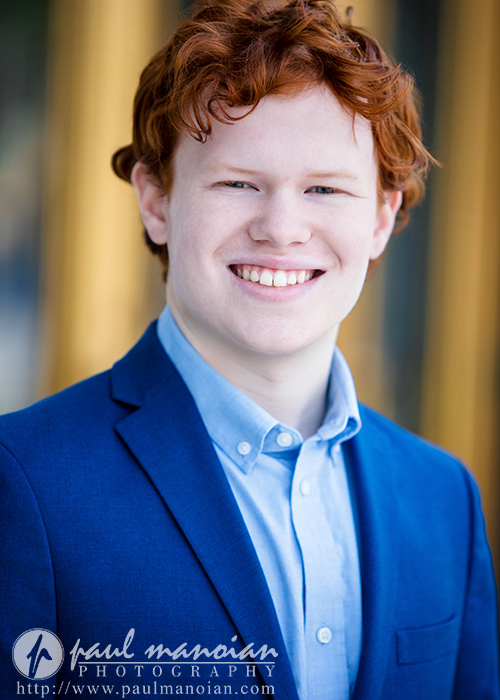Smiling high school senior with red hair in a blue suit jacket and light blue shirt, standing outdoors in Detroit, posing for Utica Academy of International Studies senior pictures.