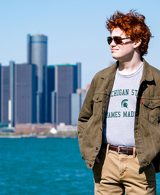 A high school senior with red hair stands by the water with a city skyline in the background on a sunny day, capturing perfect Senior Pictures for Utica Academy of International Studies.
