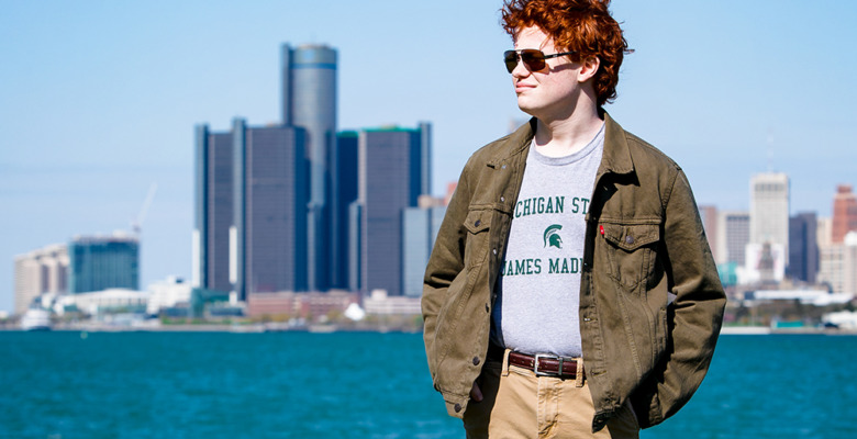 A high school senior with red hair stands by the water with a city skyline in the background on a sunny day, capturing perfect Senior Pictures for Utica Academy of International Studies.