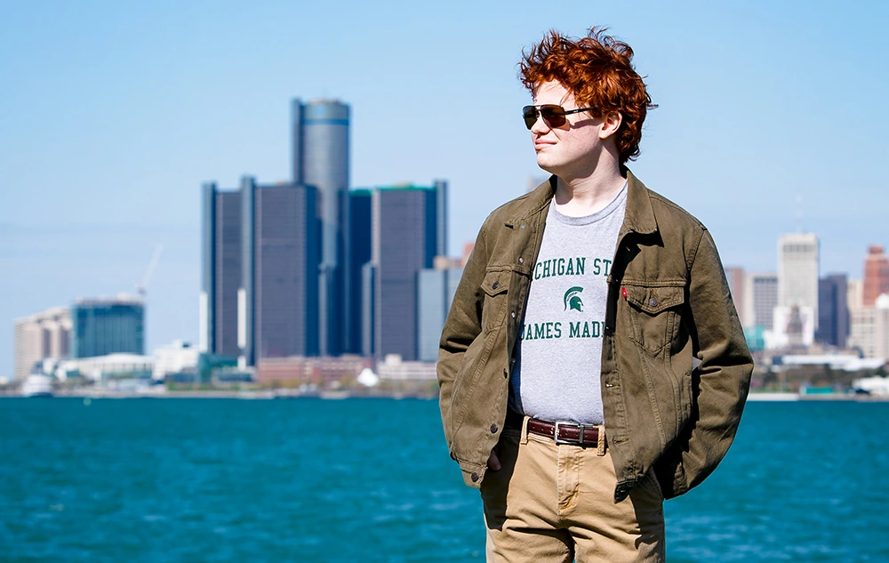 A high school senior with red hair stands by the water with a city skyline in the background on a sunny day, capturing perfect Senior Pictures for Utica Academy of International Studies.