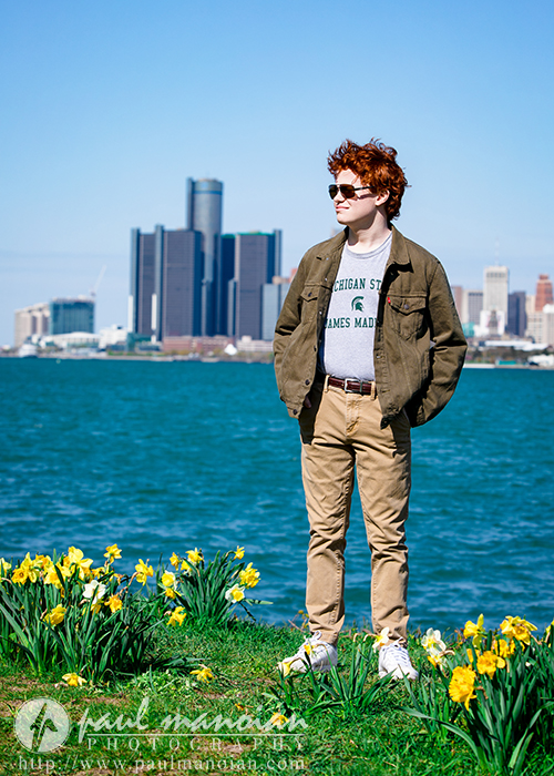 A high school senior stands by water with flowers in front and the Detroit city skyline in the background on a sunny day, taking senior pictures.