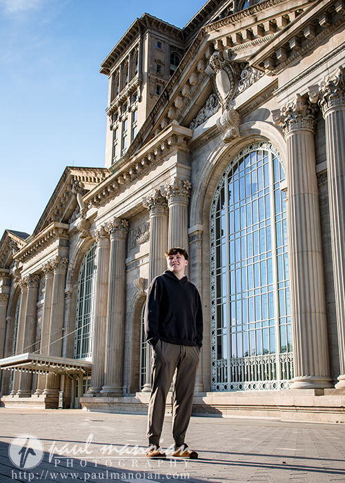 A high school senior in casual clothes stands in front of a historic building with tall columns and large arched windows during his Sunrise Session Detroit for Notre Dame Prep senior pictures.