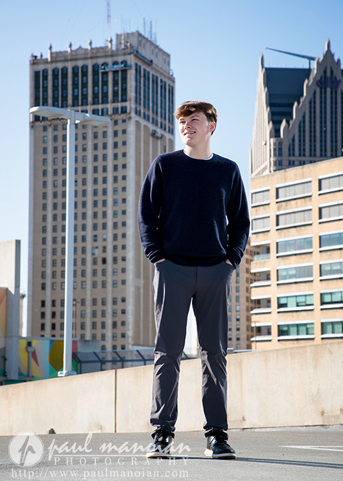 High school senior stands on a rooftop with tall city buildings in the background under a clear blue sky during his Notre Dame Prep senior pictures sunrise session.