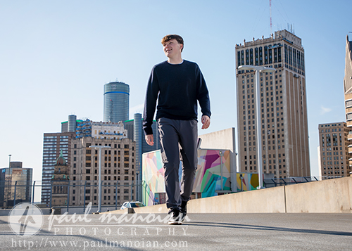 A high school senior walks on a rooftop with tall city buildings and a clear blue sky in the background, perfect for unique Senior Pictures Detroit or a stunning Sunrise Session Detroit.