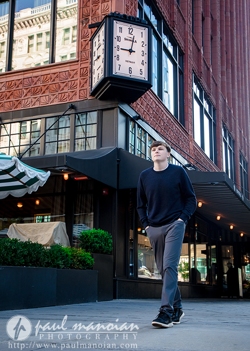 A high school senior walks on a city sidewalk beneath a vintage clock on a red brick building, capturing the spirit of Detroit senior pictures in an urban sunrise senior session.