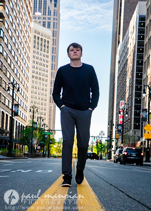 High school senior in dark clothes walks confidently down the center of a city street at sunrise, tall buildings glowing behind him—perfect for Senior Pictures Detroit or a memorable Notre Dame Prep Senior Pictures Sunrise Session.