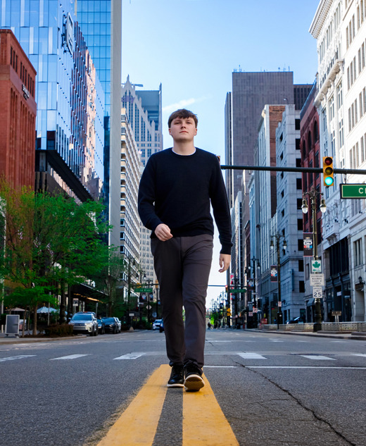 A high school senior walks down the center of a Detroit city street lined with tall buildings on a clear day, perfect for Notre Dame Prep senior pictures or a stunning sunrise session.