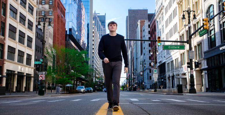 A high school senior walks down the center of a Detroit city street lined with tall buildings on a clear day, perfect for Notre Dame Prep senior pictures or a stunning sunrise session.