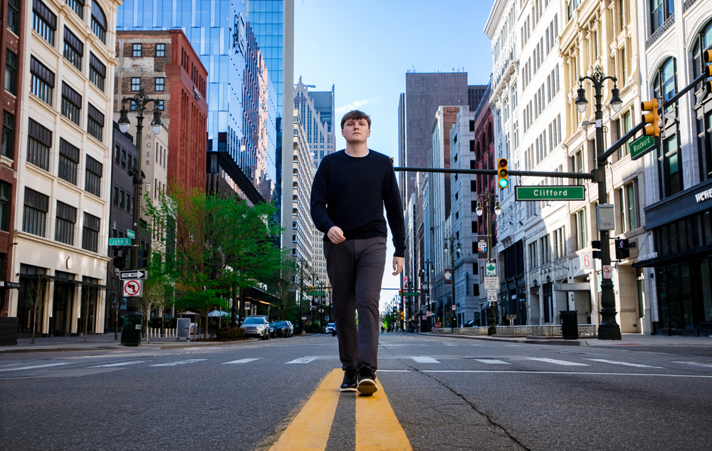 A high school senior walks down the center of a Detroit city street lined with tall buildings on a clear day, perfect for Notre Dame Prep senior pictures or a stunning sunrise session.