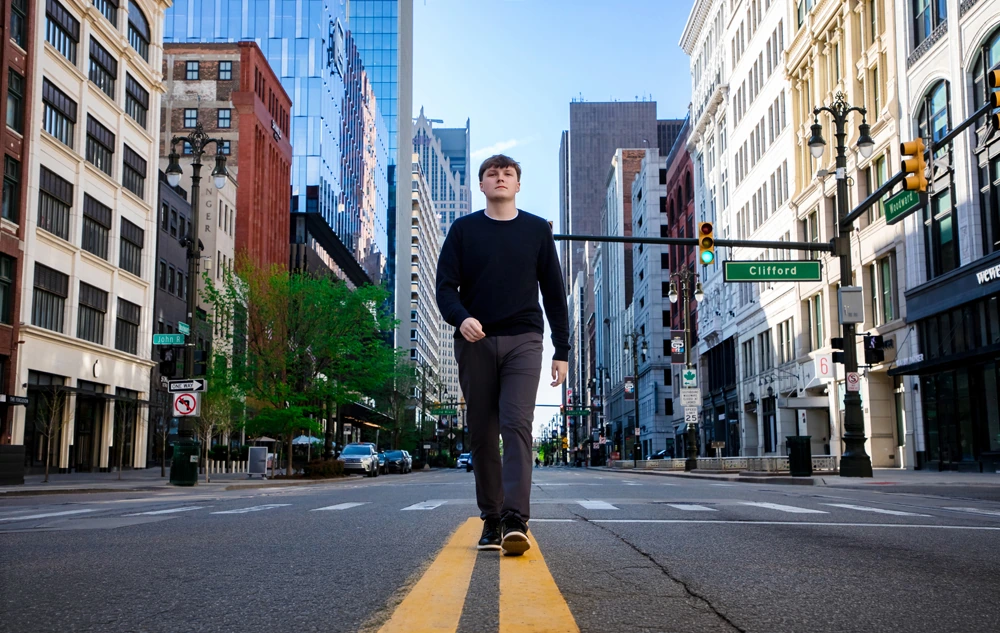 A high school senior walks down the center of a Detroit city street lined with tall buildings on a clear day, perfect for Notre Dame Prep senior pictures or a stunning sunrise session.