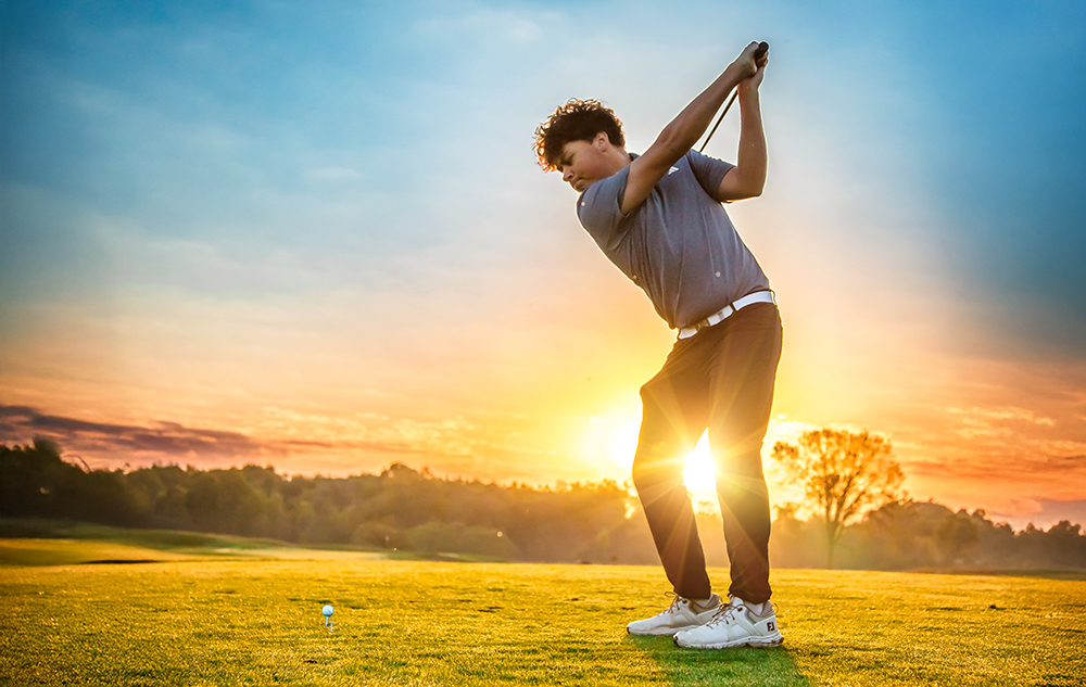 A high school senior boy prepares to swing a golf club at a golf ball on a grassy course during a stunning sunset, trees in the background—an ideal moment for a golf senior pictures session.