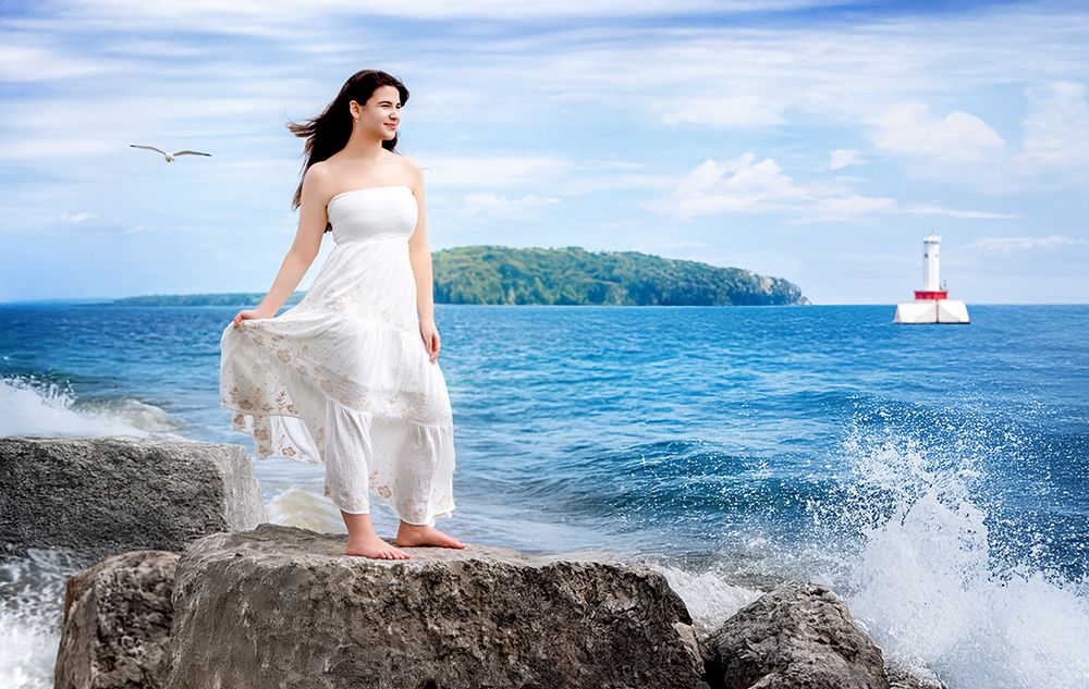 A woman in a white dress stands barefoot on rocks by the sea during a summer senior pictures session. Waves crash nearby, a seagull flies overhead, and an island with a lighthouse sits in the distance under the blue sky.