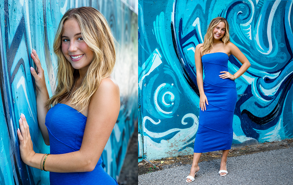 A young woman with long blonde hair wears a strapless blue dress and white sandals, smiling while posing in front of a vibrant blue and turquoise graffiti mural.