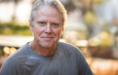 Middle-aged man with short gray hair and a light gray long-sleeve shirt smiles softly at the camera outdoors, with a blurred background of greenery and warm sunlight—captured by an Ann Arbor Actor Headshot Photographer.