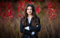 A young woman with long brown hair, wearing a black suit and white shirt, stands in tall grass surrounded by red and green foliage—a classic fall senior pictures session. She faces forward with arms crossed and a slight smile.