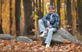 A young person in a plaid shirt and jeans sits on a large rock in a forest, surrounded by autumn leaves—perfect for a fall senior pictures session. Trees with golden foliage form a beautiful background.