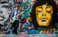 A young person stands against a colorful graffiti wall, playing an electric guitar. The wall features large painted faces and vibrant, multicolored designs. The person wears glasses, a plaid shirt, and dark pants.