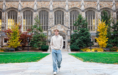 A young person in a light sweater and jeans walks on a stone path in front of a large, historic building with tall arched windows, autumn trees on either side—a perfect spot for a fall senior pictures session.