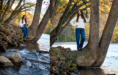 A young woman with long brown hair, wearing a white sweater and blue jeans, poses on the roots of a large tree by a riverbank, surrounded by autumn foliage during her fall senior pictures session.