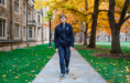 A person wearing dark clothing walks on a stone path through a campus with historic buildings and autumn trees—an ideal setting for a fall senior pictures session, with green grass and scattered fallen leaves enhancing the scene.