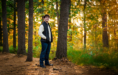 A young man with curly hair, glasses, and a black vest over a light sweater stands in a sunlit forest, smiling with his hands in his pockets during his fall senior pictures session. Tall trees and green foliage surround him as sunlight filters through.