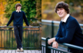 A young man with curly brown hair, wearing a navy sweater, white collared shirt, and dark pants, stands and leans on a metal railing outdoors, smiling during his fall senior pictures session with autumn foliage in the background.