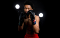 A young male boxer with short curly hair poses in a boxing stance, wearing black gloves and red shorts—perfect for dynamic boxing senior pictures—framed by two bright studio lights against a dark background.