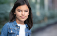 A young girl with straight dark hair and brown eyes looks at the camera, wearing a blue denim jacket over a white shirt, standing outdoors with a blurred green background.