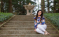 A woman with long dark hair, wearing a colorful floral blouse and white pants, sits smiling on stone steps outdoors, surrounded by greenery and trees, with a blurred stone fountain in the background.