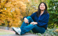 A young woman with long dark hair, wearing a blue sweater and jeans, sits on a stone ledge outdoors. She is smiling and surrounded by autumn trees with yellow and green leaves.