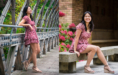 A young woman in a mauve floral dress and wedge sandals poses outdoors; on the left, she stands on a wooden bridge, and on the right, she sits on a stone bench near pink flowers, smiling in both scenes.