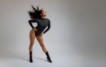 Woman in a black bodysuit and boots poses playfully with hair flipping in a studio with a grey background.
