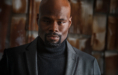 A man with a shaved head, goatee, and mustache wears a black turtleneck and gray blazer, standing confidently before brown, textured panels—an ideal example of how to prepare for actor headshots with style and poise.