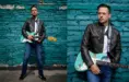 A Detroit musician in a leather jacket and jeans stands against a turquoise brick wall, holding a weathered electric guitar. He appears focused, with a checkered strap across his shoulder, exuding a relaxed, musical vibe that would captivate any Detroit music photographer.