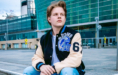 High school senior guy wearing a Detroit Lions varsity jacket with the number 65, sitting in front of the Renaissance Center building in downtown Detroit, MI for his senior pictures session with award-winning senior photographer, Paul Manoian. He has light brown hair and are looking to the side, with a thoughtful expression.