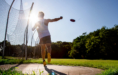 A high school senior throws a discus during a sunny outdoor practice, with the sun shining brightly behind them and trees in the background. The athlete is in mid-motion inside a throwing circle.