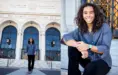 Smiling high schools senior with long, curly hair standing and sitting in front of the Detroit Institute of Arts entrance for his downtown Detroit senior pictures session with award-winning Detroit photographer, Paul Manoian. They're wearing a blue-gray shirt and black pants. The building's grand architecture features large arched windows and decorative details.