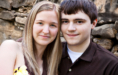 A young couple stands close together, smiling softly at the camera in front of a stone wall—an intimate moment captured as part of their wedding engagement portraits. The woman has straight blonde hair; the man, short brown hair and a dark polo shirt.