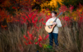A person wearing a cream sweater stands in tall grass and red autumn foliage, holding an acoustic guitar during a fall senior pictures session. Bright yellow leaves in the background complete this vibrant autumn scene.