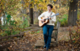 A person wearing glasses and a cream sweater plays an acoustic guitar while standing on a wooden path in a forest, surrounded by autumn leaves—perfect for a cozy fall senior pictures session.