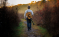 A person wearing a white sweater and jeans walks down a narrow, grassy path through autumnal brush during a fall senior pictures session, carrying an acoustic guitar on their back, framed by golden light in the background.